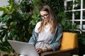 Woman gardener sitting on chair in green house, working on laptop surrounded by plants, remote work, distance job Royalty Free Stock Photo