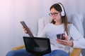 Woman in glasses and headphones is working on laptop sitting in armchair. Mobile phone and tablet in hands.