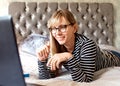 Woman in glasses lying on a bed looking at a laptop and smiling