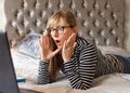 Woman in glasses lying on a bed looking at laptop and wondering