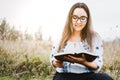Woman with glasses sitting outdoor on the grass holding and reading an open Bible. Sunlight Royalty Free Stock Photo