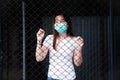 Woman in a green medical face mask in a white shirt stands behind a black fence  showing a sad expression. Royalty Free Stock Photo