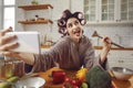 Woman in hair curlers taking funny selfie on mobile while cooking in the kitchen Royalty Free Stock Photo