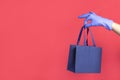 Woman hand in glove with gift paper bag on red background. Delivery of food, groceries, home shopping. Protection for COVID-19