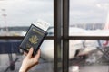 Woman hand holding american passport with boarding pass in the airport waiting zone looking through the window at planes.