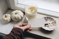 Woman hand holding blank business card mockup. Burning candle, matches, old books on windowsill. Winter, autumn still