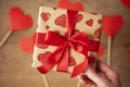 Woman hand holding gift box with red bow. Many hearts on a wooden background in defocus. Valentine`s Day concept Royalty Free Stock Photo