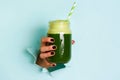 Woman hand holding glass jar of green smoothie, fresh juice against blue background. Healthy beverage, vegan, vegetarian concept. Royalty Free Stock Photo