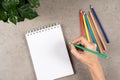 Woman hand holding green colored pencil over blank white recycled paper spiral notebook on gray desk background. Top
