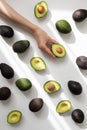 Woman hand holding half avocado of heap whole avocados on table top view in aesthetic shadow sunlight. Flat lay