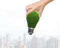 Woman hand holding one light bulb with green grass, on city buildings skyscrapers background, concept of ECO and green energy Royalty Free Stock Photo