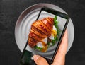 Woman hand holding and showing smart phone takes a photo Croissant sandwich with poached egg, tomato and avocado on plate on Dark Royalty Free Stock Photo