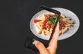 Woman hand holding and showing smart phone takes a photo Pasta penne bolognese in white plate on wooden table background