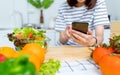 Woman hand holding a smartphone and salad bowl with tomato and various green leafy vegetables on the table at the home.