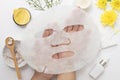 Woman hand holding white sheet mask with clay mask wooden spoon yellow flowers and serum skincare cream products on white