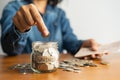 The woman hand is putting a coin in a glass bottle and a pile of coins on a brown wooden table,Investment business, retirement, Royalty Free Stock Photo