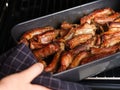 A woman hand taking a black baking tray with baked pork ribs out of an oven