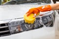 Woman hand wearing orange gloves with yellow sponge washing headlight modern car or cleaning automobile.