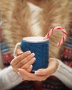 Woman hands with elegant french manicure nails design holding a cozy knitted mug with hot cocoa or coffee with a mint candy cane.
