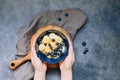 Woman hands hold Dark blue bowl of oatmeal porridge with banana and blueberry on vintage table top view in flat lay