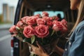 Woman holding bouquet of tender pink roses and is about to put them in the trunk of a cargo minibus. Flowers delivery