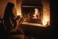 Woman hands holding cup of tea and warming up at cozy fireplace in dark evening room, close up. Fireplace heating in house,