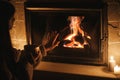 Woman hands holding cup of tea and warming up at cozy fireplace in dark evening room, close up. Fireplace heating in house,