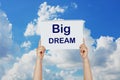 Woman hands holding high a post card that says BIG DREAM against a blue sky