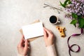 Woman hands holding kraft paper envelope with blank card over feminine desk decorated with coffee cup, cookies, lilac flowers and