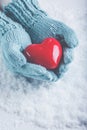 Woman hands in light teal knitted mittens are holding beautiful glossy red heart in snow background. Love, St. Valentine concept