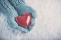 Woman hands in light teal knitted mittens are holding beautiful glossy red heart on snow background. Love, St. Valentine concept