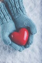 Woman hands in light teal knitted mittens are holding beautiful glossy red heart in snow background. Love, St. Valentine concept