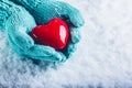 Woman hands in light teal knitted mittens are holding a beautiful glossy red heart in a snow background. St. Valentine concept.