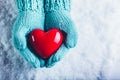 Woman hands in light teal knitted mittens are holding a beautiful glossy red heart in a snow background. St. Valentine concept. Royalty Free Stock Photo