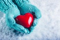 Woman hands in light teal knitted mittens are holding a beautiful glossy red heart in a snow background. St. Valentine concept.