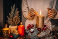 Woman hands lightning big gold candle with matches in Christmas wreath. Xmas interior decoration on rustic wooden background. hygg Royalty Free Stock Photo