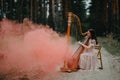 Woman harpist sits at forest and plays harp against a background of pines and smoke