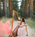 Woman harpist sits at forest and plays harp against a background of pines and smoke