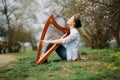 Woman harpist sits on grass and plays harp among blooming apricot trees Royalty Free Stock Photo