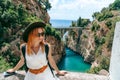 A woman in a hat with a backpack enjoys an incredibly beautiful view of a stone bridge over the gorge Fiordo di Furore. Little bay