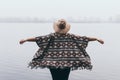 Woman in hat and poncho standing on the river bank and looking towards autumn forest covered with morning fog Royalty Free Stock Photo