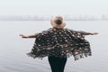 Woman in hat and poncho standing on the river bank and looking towards autumn forest covered with morning fog Royalty Free Stock Photo