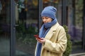 Woman in hat and scarf in beige coat sending text message using smartphone outside in city in winter Royalty Free Stock Photo