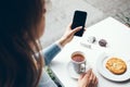 Woman is having breakfast in a cafe, tea with cookie and is using internet on her smart phone, reading news, checking e-mail and