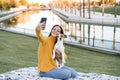 woman having fun with jack russell dog in park, sitting on blanket during autumn season. Woman taking picture with mobile phone. Royalty Free Stock Photo