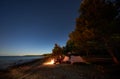 Woman having a rest at night camping near tourist tent, campfire on sea shore under starry sky