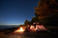 Woman having a rest at night camping near tourist tent, campfire on sea shore under starry sky Royalty Free Stock Photo