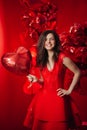 Woman with Hearts Balloons on a red background. Beauty Girl on Valentine`s Day. Model in a stylish red dress