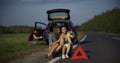 Two girls sit on the spare tire and take a selfie while a man fixes something in a broken car. emergency stop sign