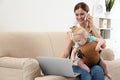 Woman with her son in baby carrier using laptop and talking on phone at home. Royalty Free Stock Photo